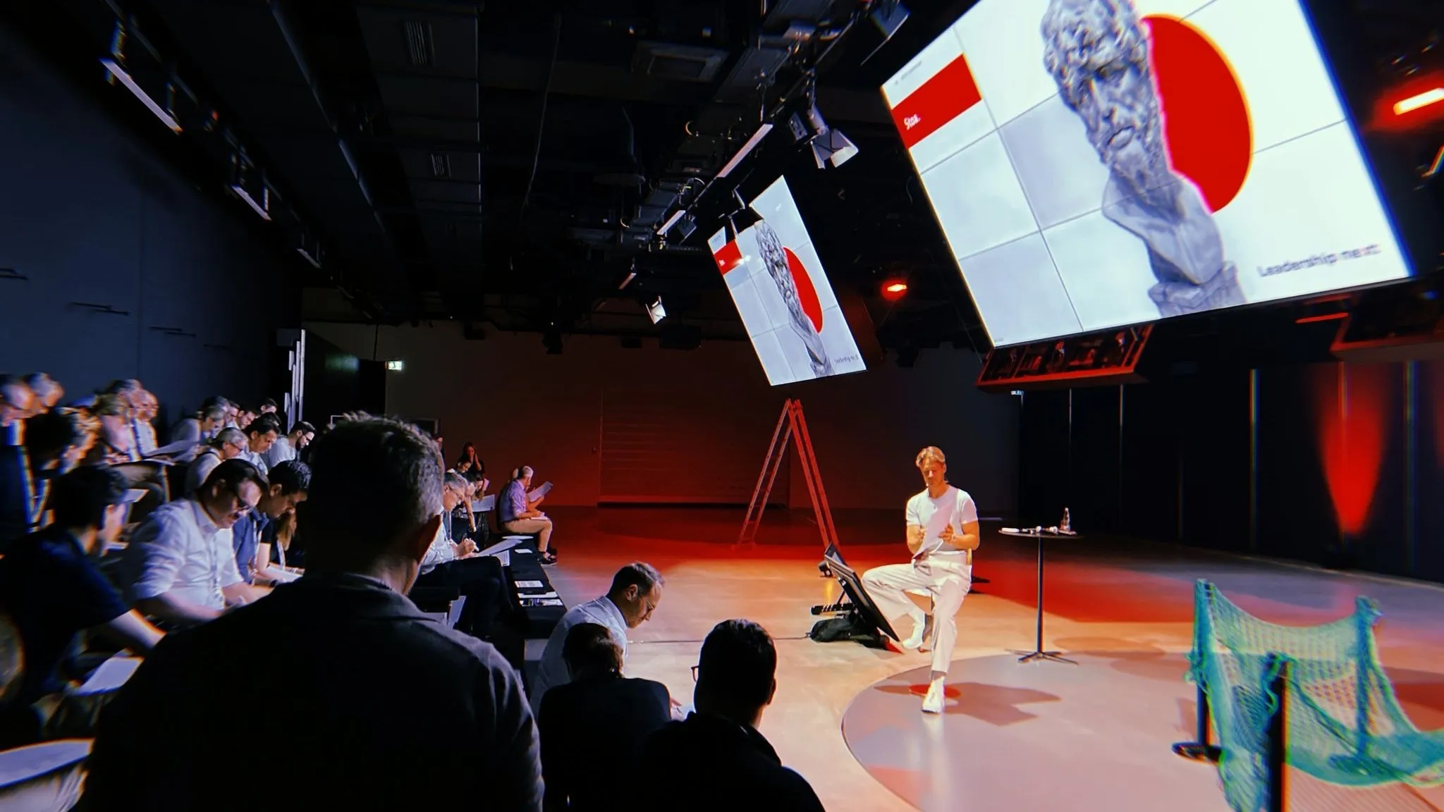 Führungskräfte im abgedunkelten Studio, auf der Bühne ein Facilitator vor zwei großen Leinwänden mit der Kampagnengrafik „Leadership next“
