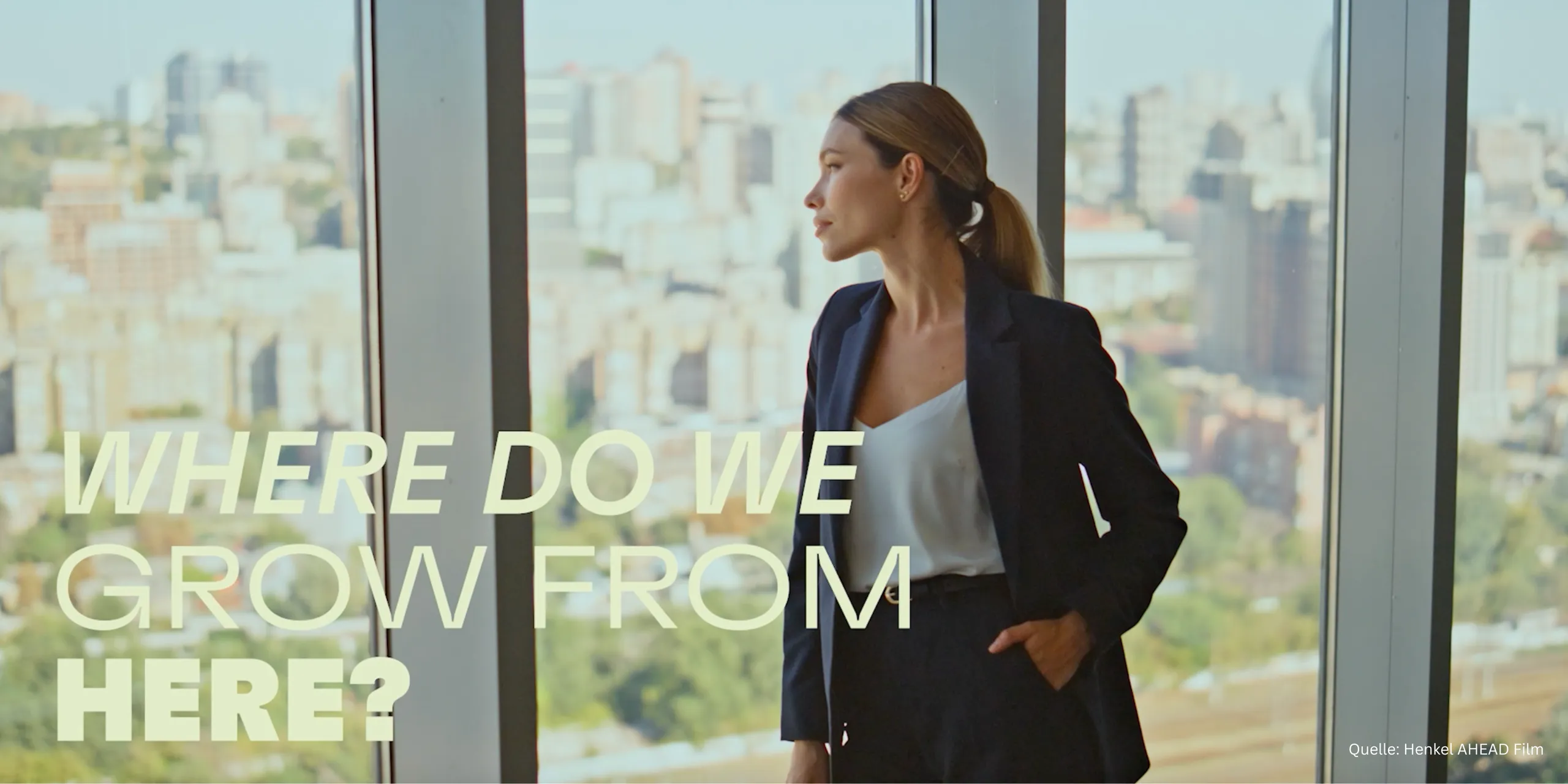 Filmstill aus dem Henkel-AHEAD-Film: eine Frau im dunklen Anzug blickt aus einem Hochhausfenster über eine Großstadt, darüber die Zeile „Where do we grow from here?“