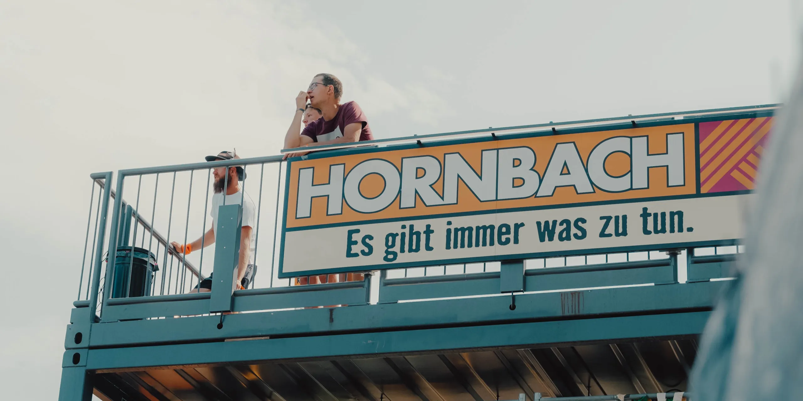 Drei Menschen stehen auf dem Dach eines HORNBACH-Marktes hinter dem Werbeschild „HORNBACH – Es gibt immer was zu tun.“ und blicken in die Ferne